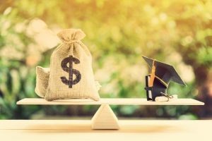 Money cost saving for goal and success in school, education concept : US dollar bills / cash in burlap bags, a black graduation cap or hat, a certificate / diploma and a book on basic balance scale.