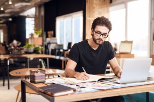 Freelancer bearded man taking notes at laptop sitting at desk.