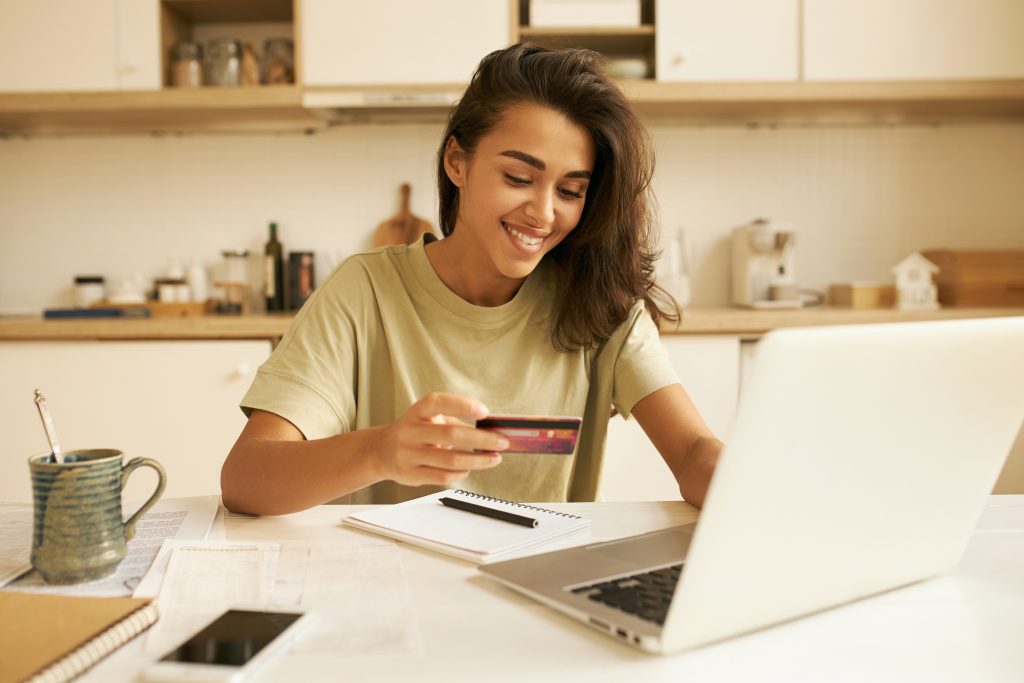 Stylish pretty student girl surfing internet in kitchen, sitting at table with open laptop and mug, holding plastic credit card, entering information via paying system while shopping online